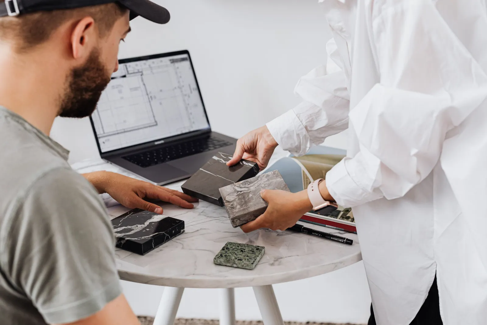 Designer and homeowner at a table comparing stone samples beside a laptop showing a floor plan