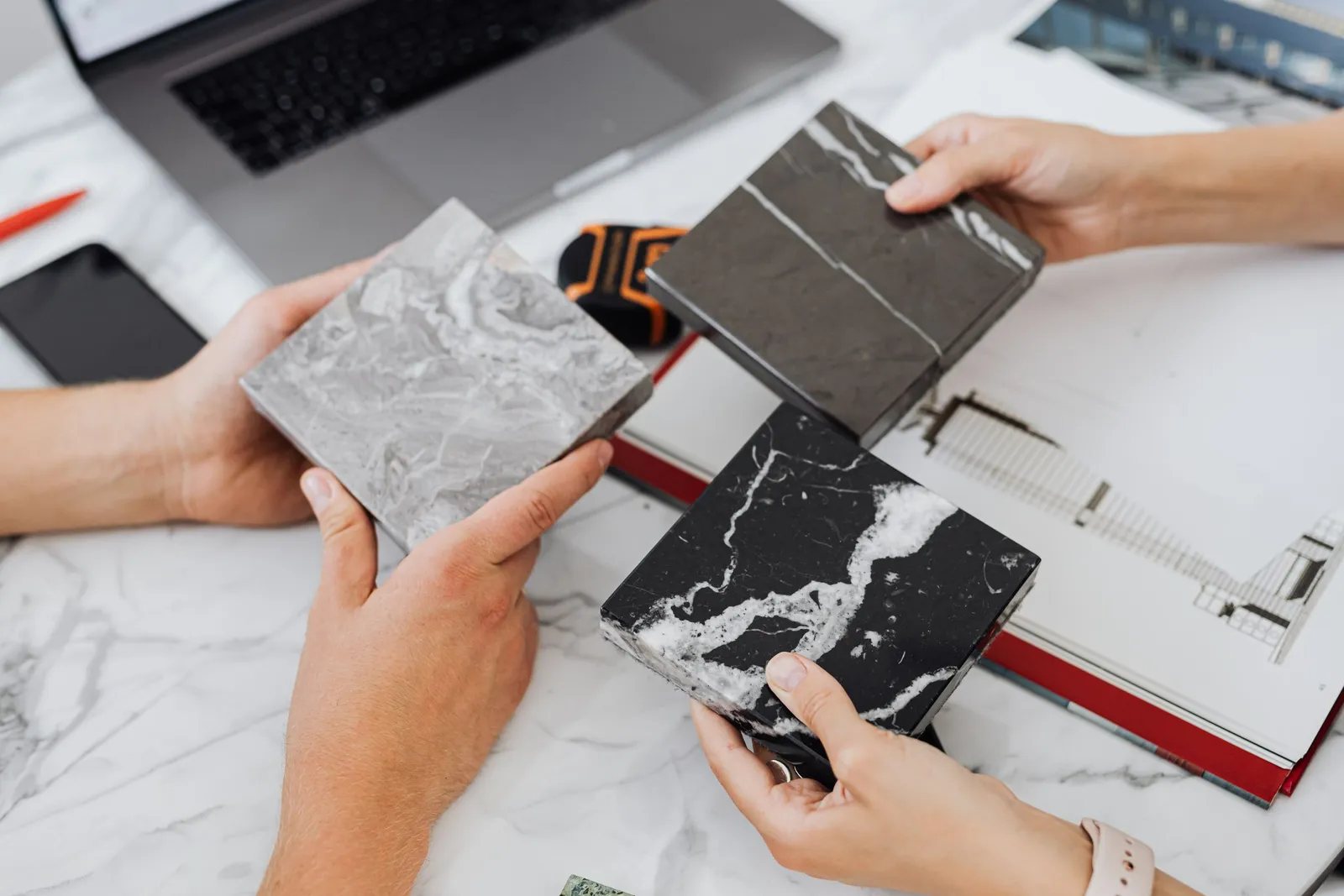 Hands holding four marble and stone tile samples over a desk with a laptop